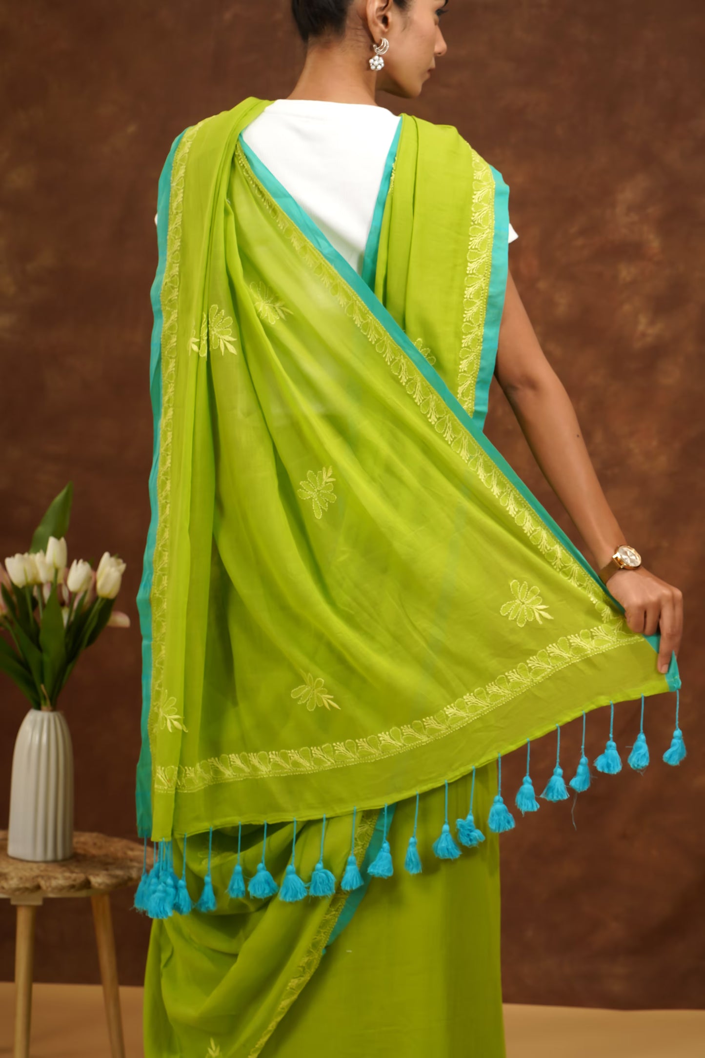 Woman wearing a parrot green saree with blue border and tassels against a brown background