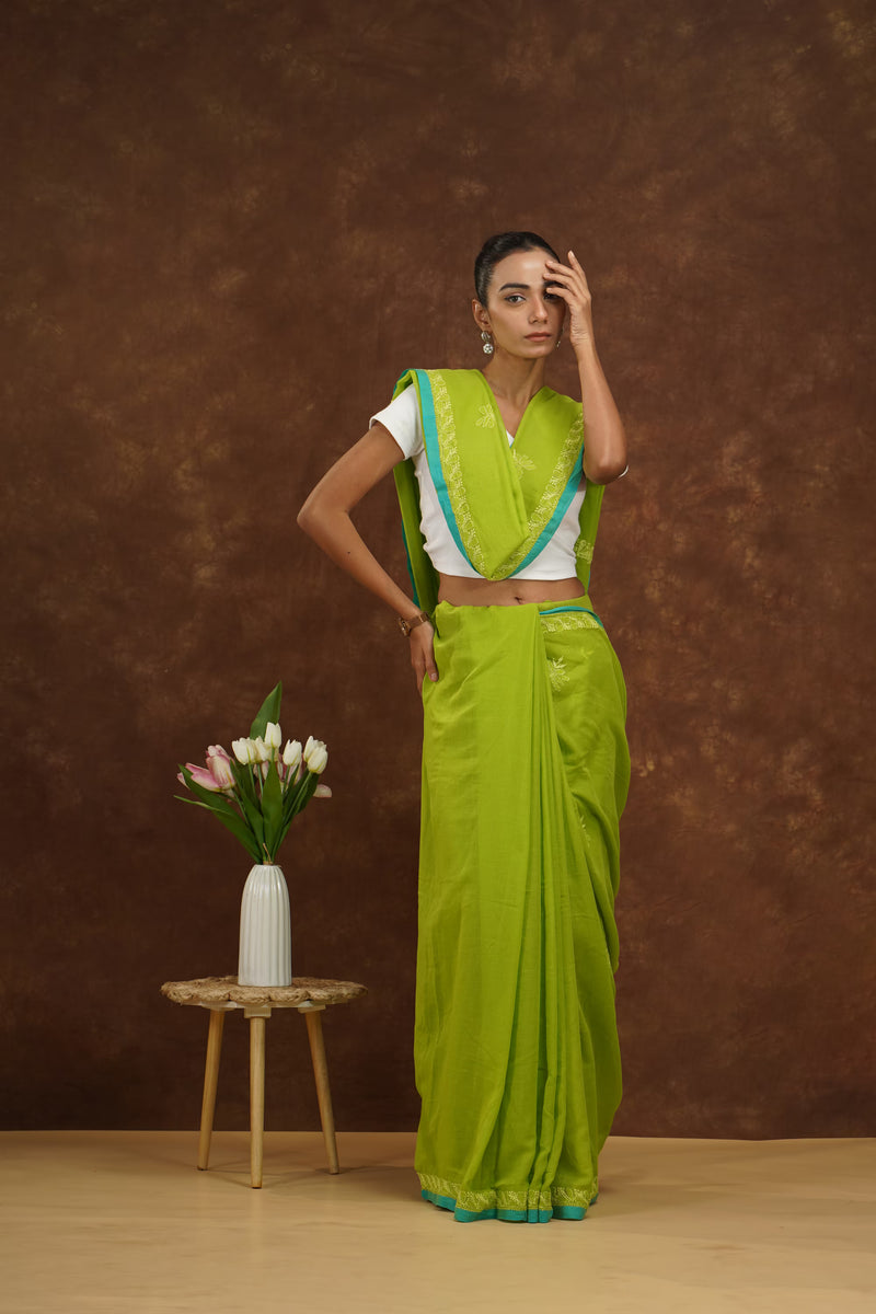 Woman in a parrot green saree standing next to a small table with flowers against a brown background