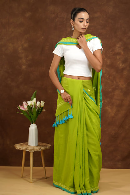 Woman in a parrot green saree with white blouse standing next to a small table with flowers against a brown background