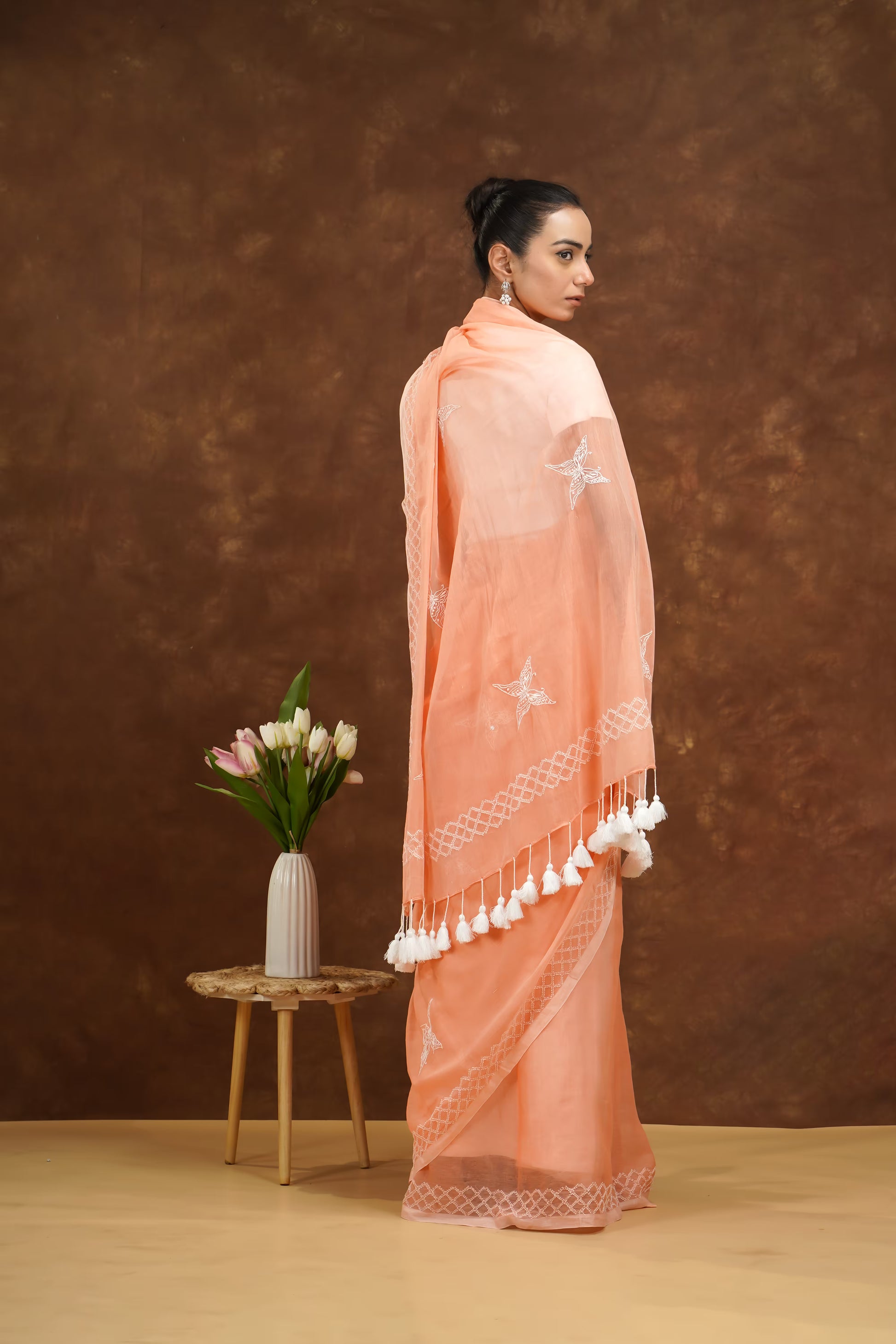 Woman wearing a peach saree with white patterns and tassels, standing against a brown background.
