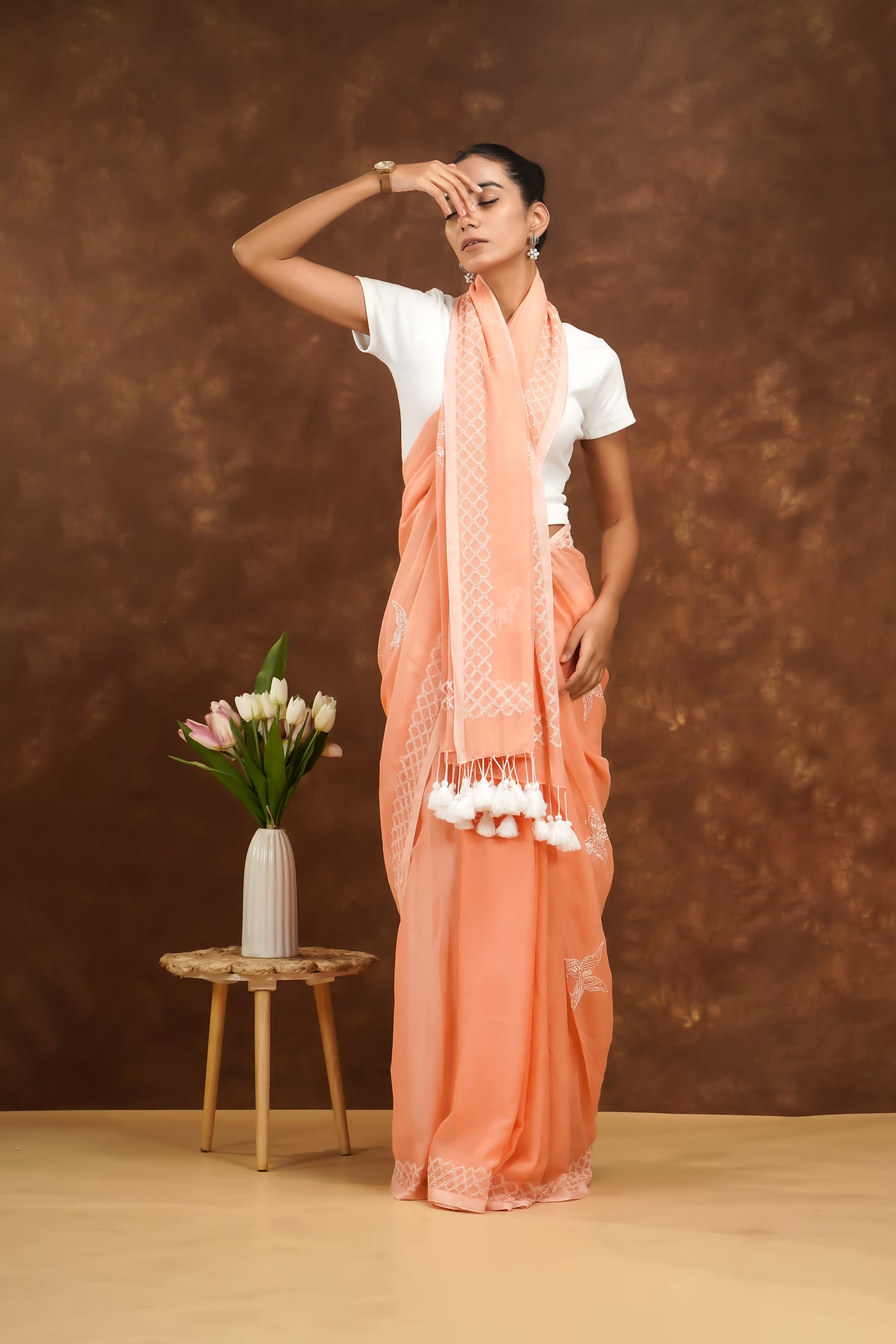 Woman in a peach saree with white patterns and tassels, standing against a brown background.