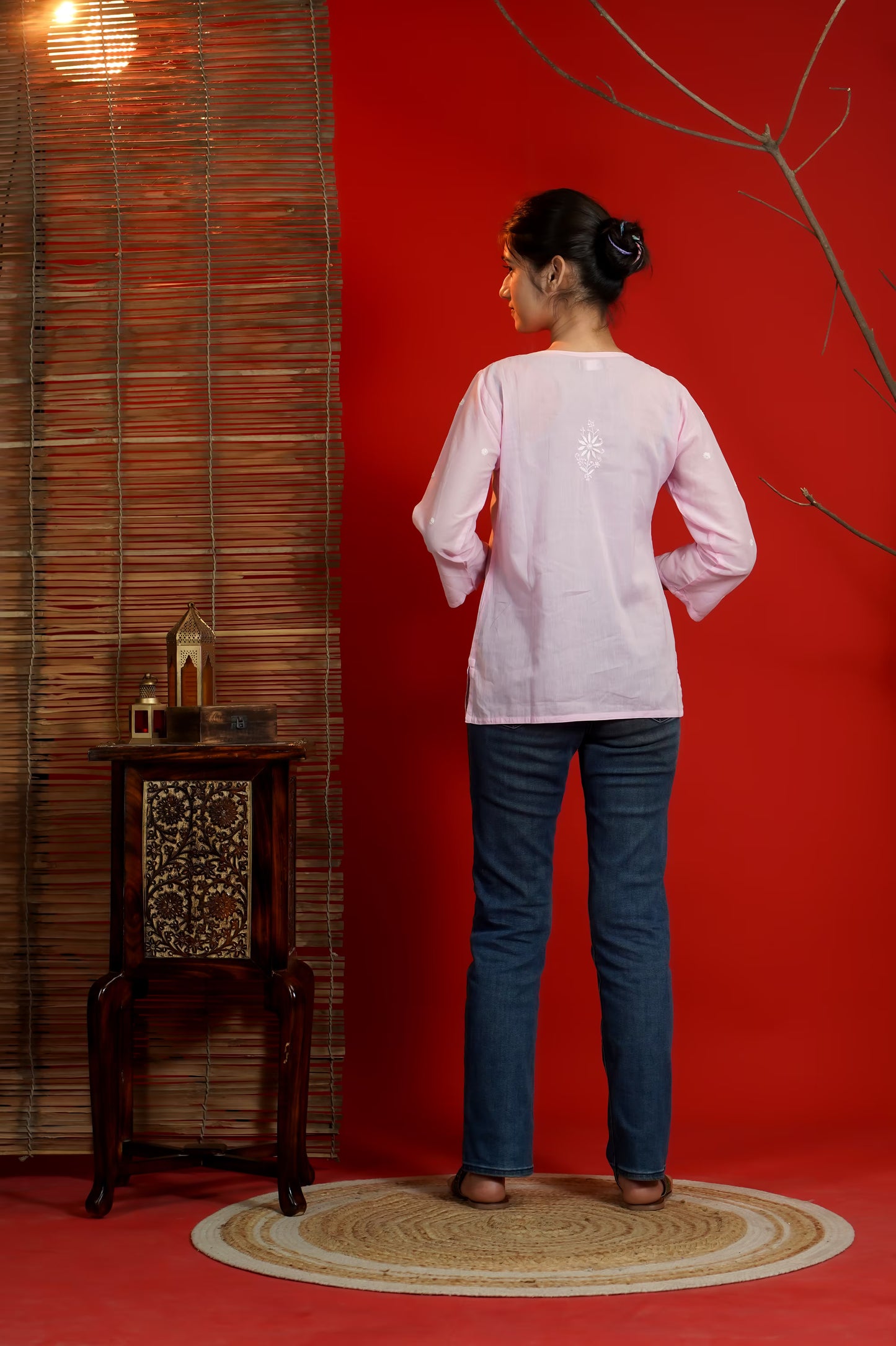 Person wearing a light pink shirt and blue jeans standing against a red wall with a wooden side table and decorative items.