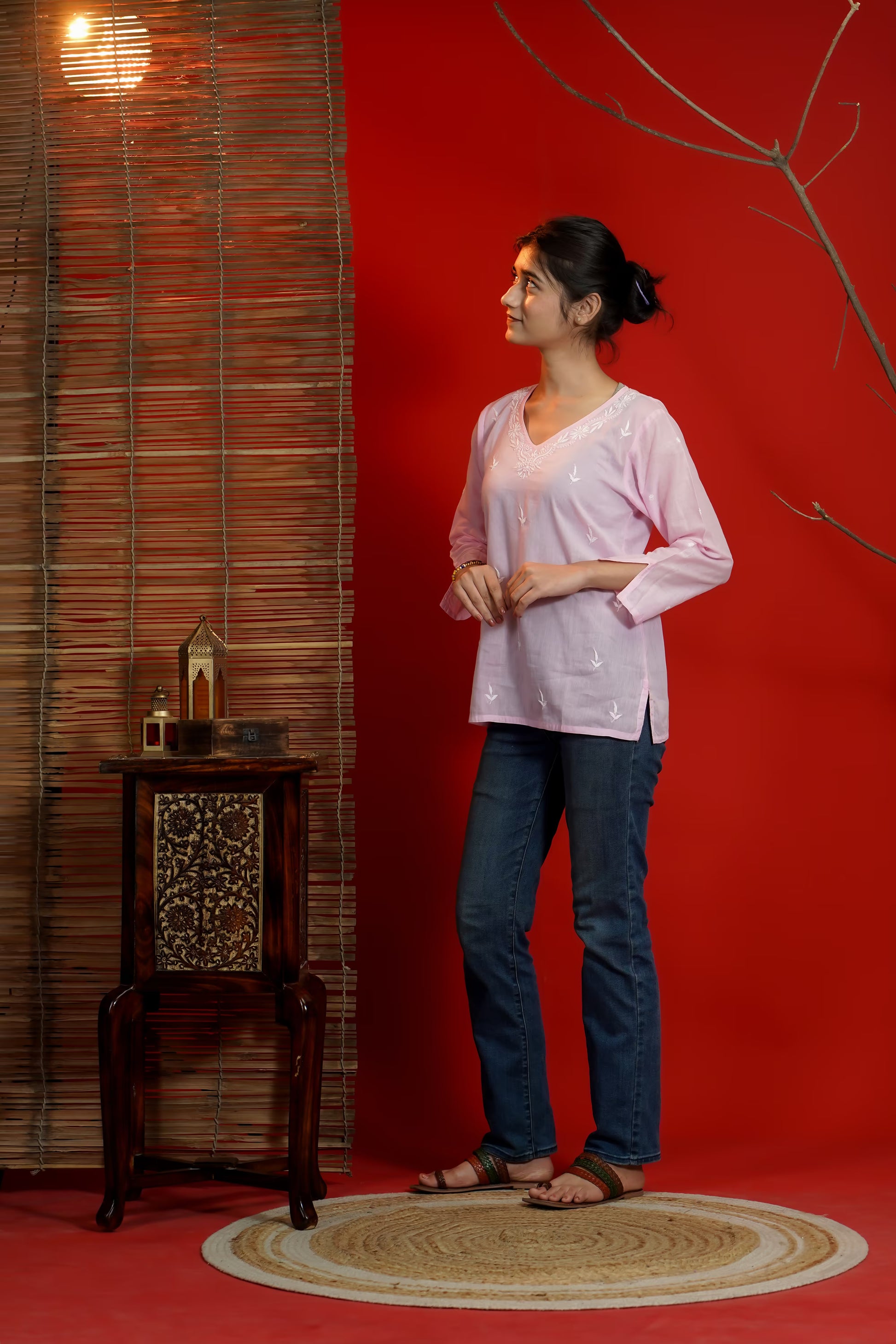 Woman in a pink top and blue jeans standing against a red wall with a wooden side table.