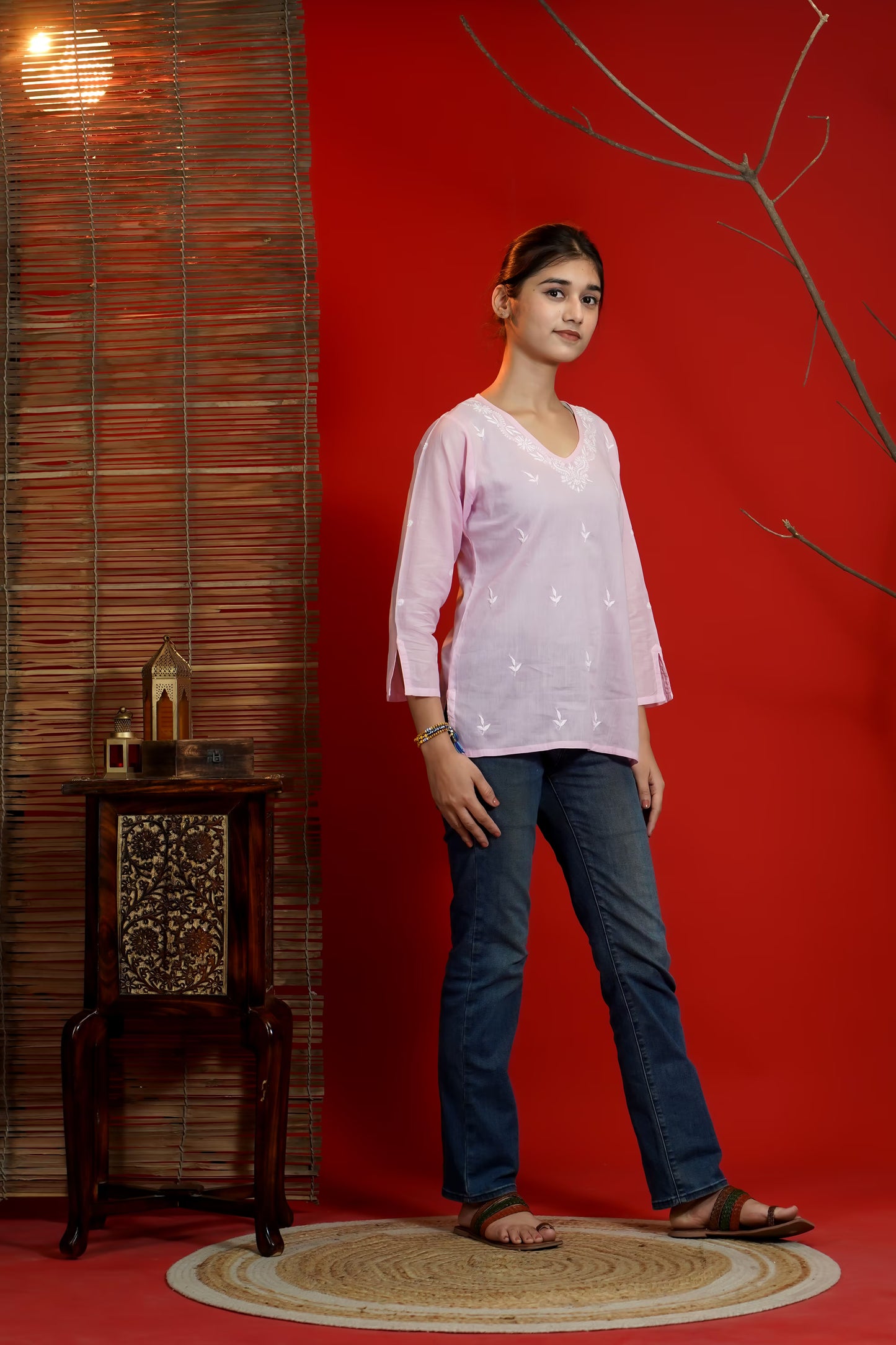 Woman in a light pink top and blue jeans standing against a red wall with a wooden side table.