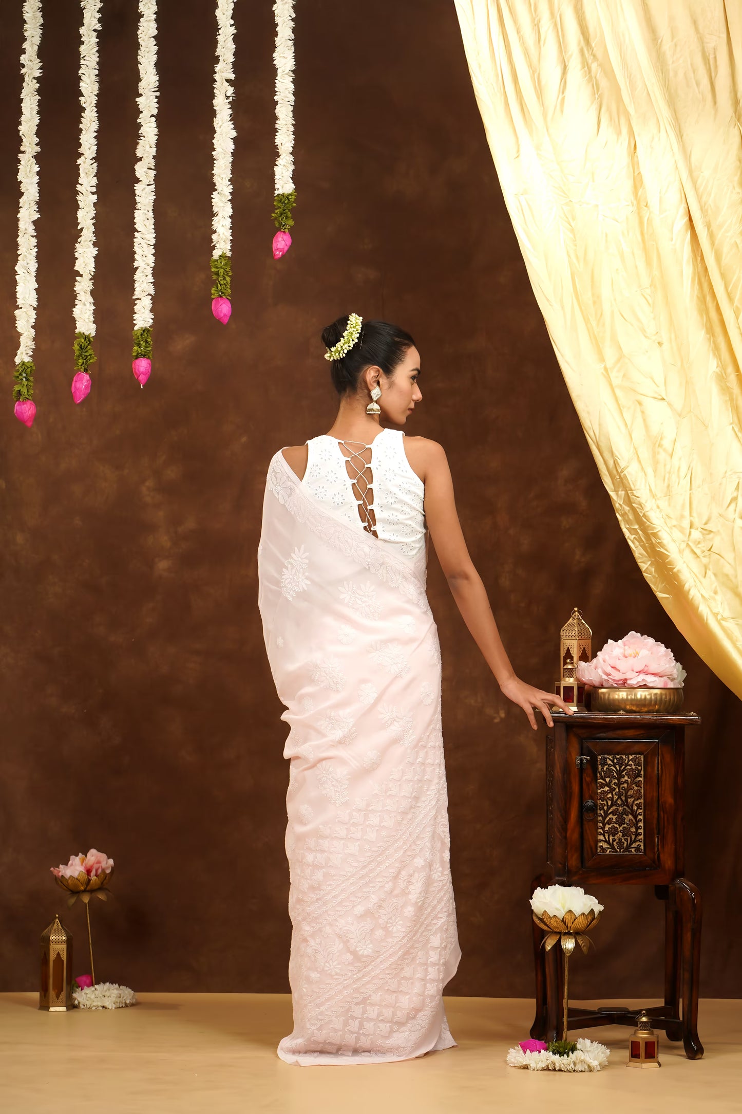 Woman in a white saree standing in front of a decorative setup with flowers and a curtain.