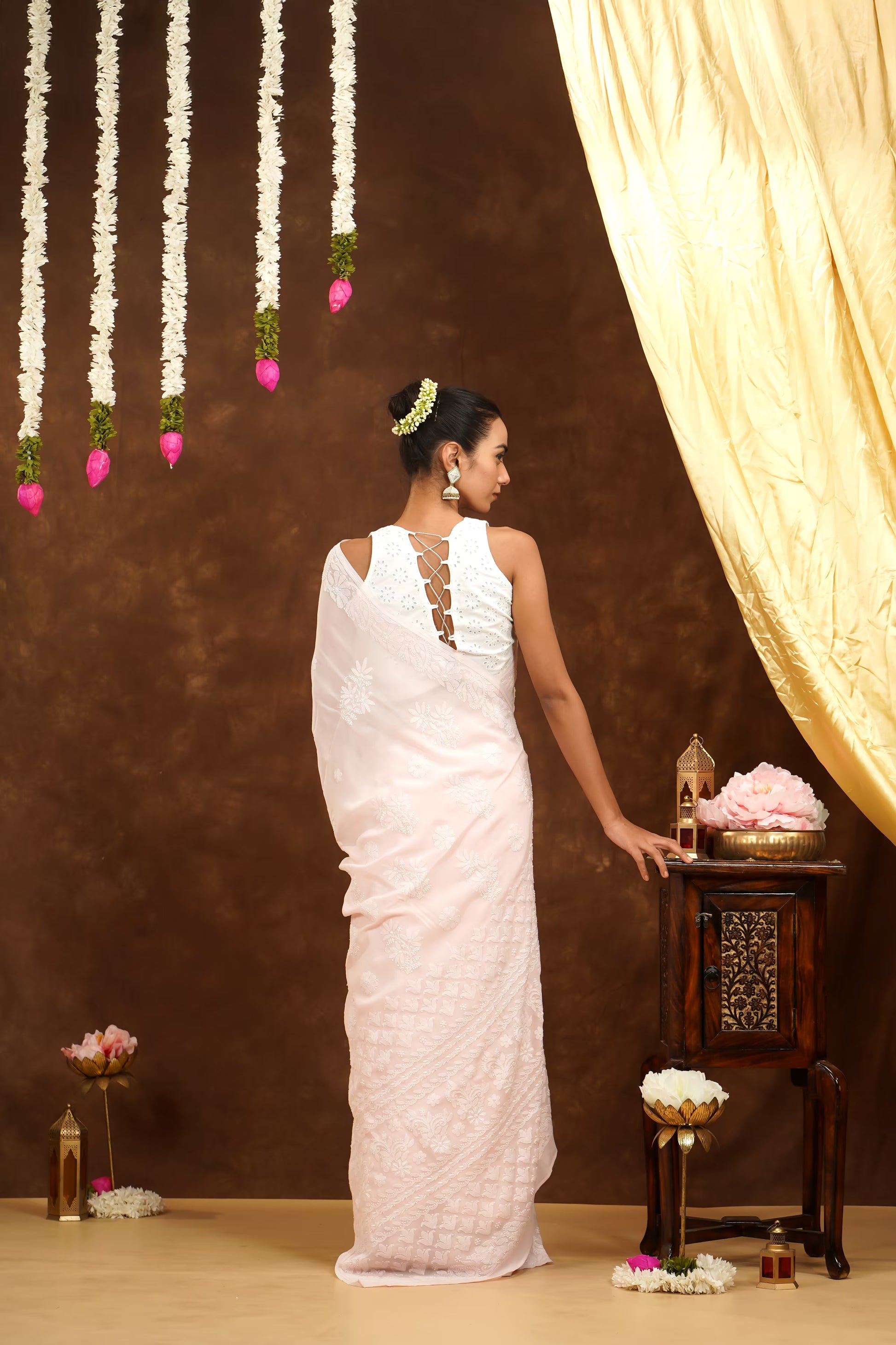 Woman in a white saree standing in front of a decorative setup with flowers and a curtain.