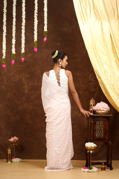 Woman in a white saree standing in front of a decorative setup with flowers and a curtain.