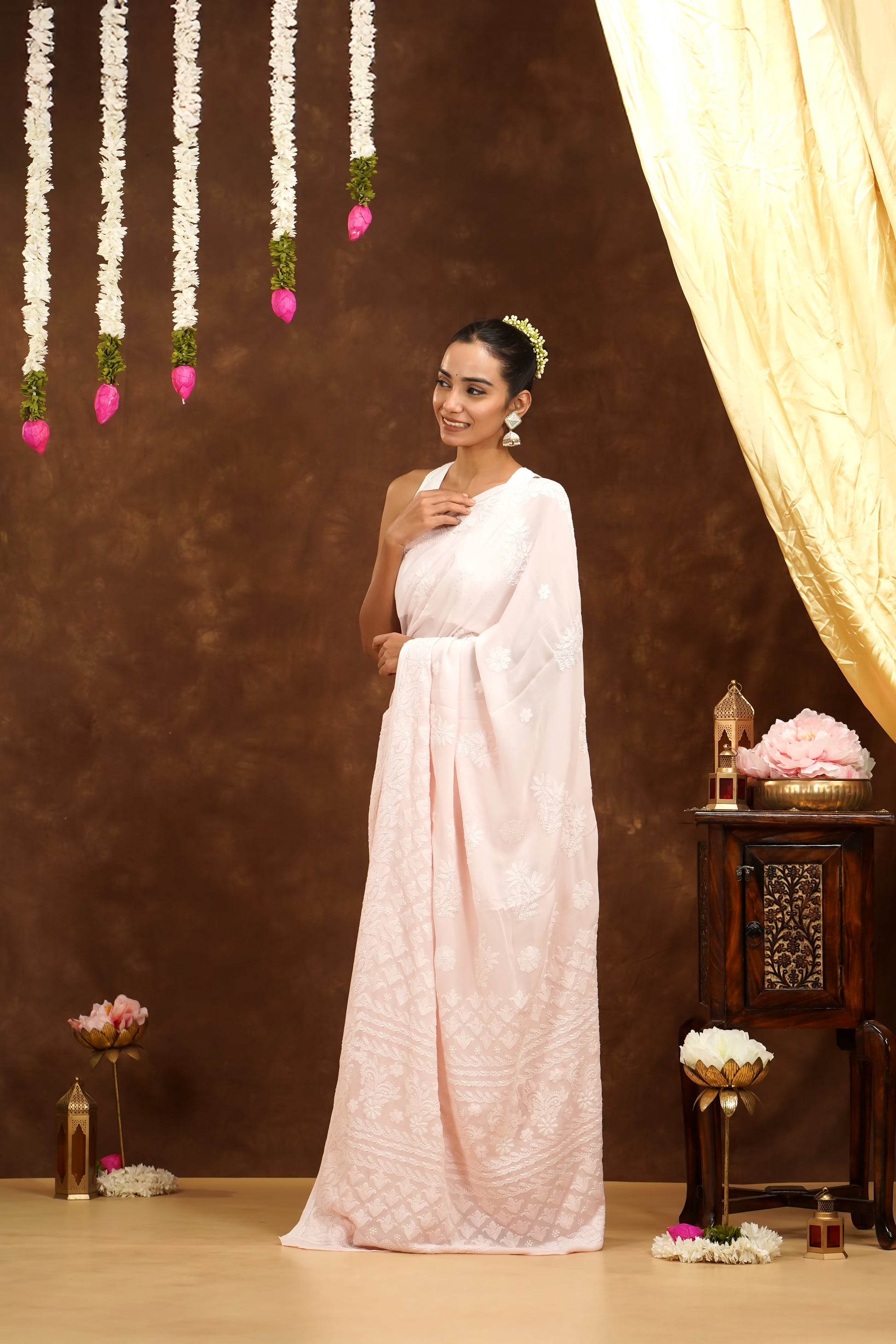 Woman in a white saree standing in a decorated room with flowers and a curtain.