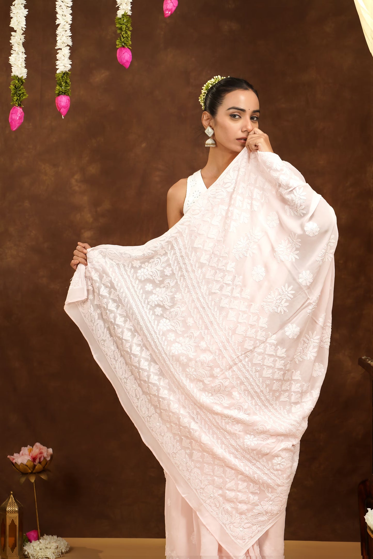 Woman holding a white embroidered saree against a brown background with decorative elements.