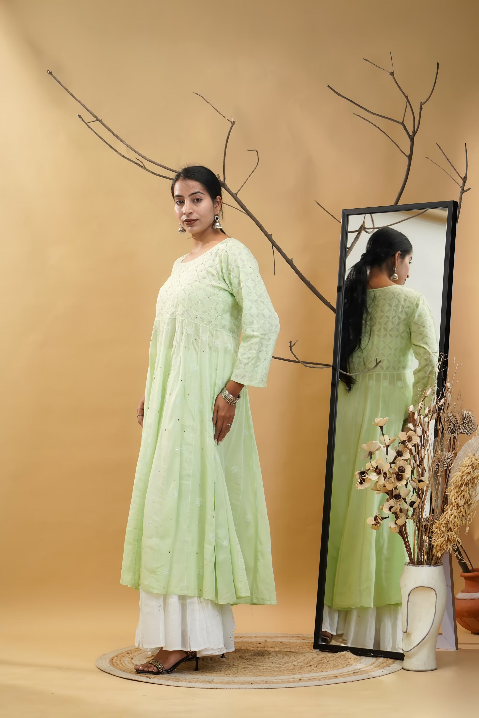 Woman in a light green traditional outfit standing next to a mirror on a beige background