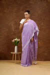 Woman in a purple saree standing next to a small table with flowers against a brown background