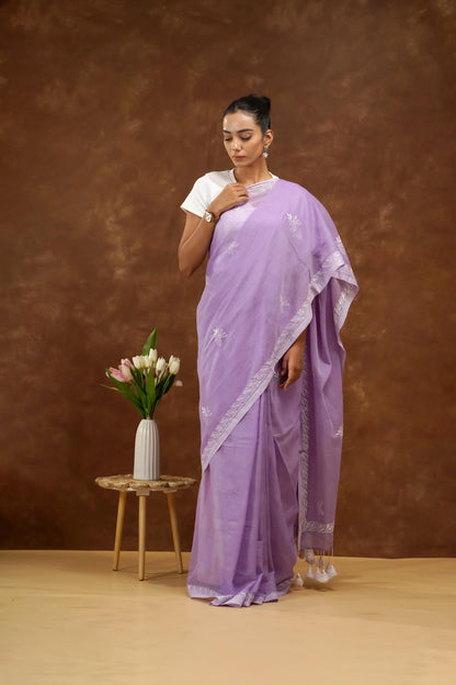 Woman in a purple saree standing next to a small table with flowers against a brown background
