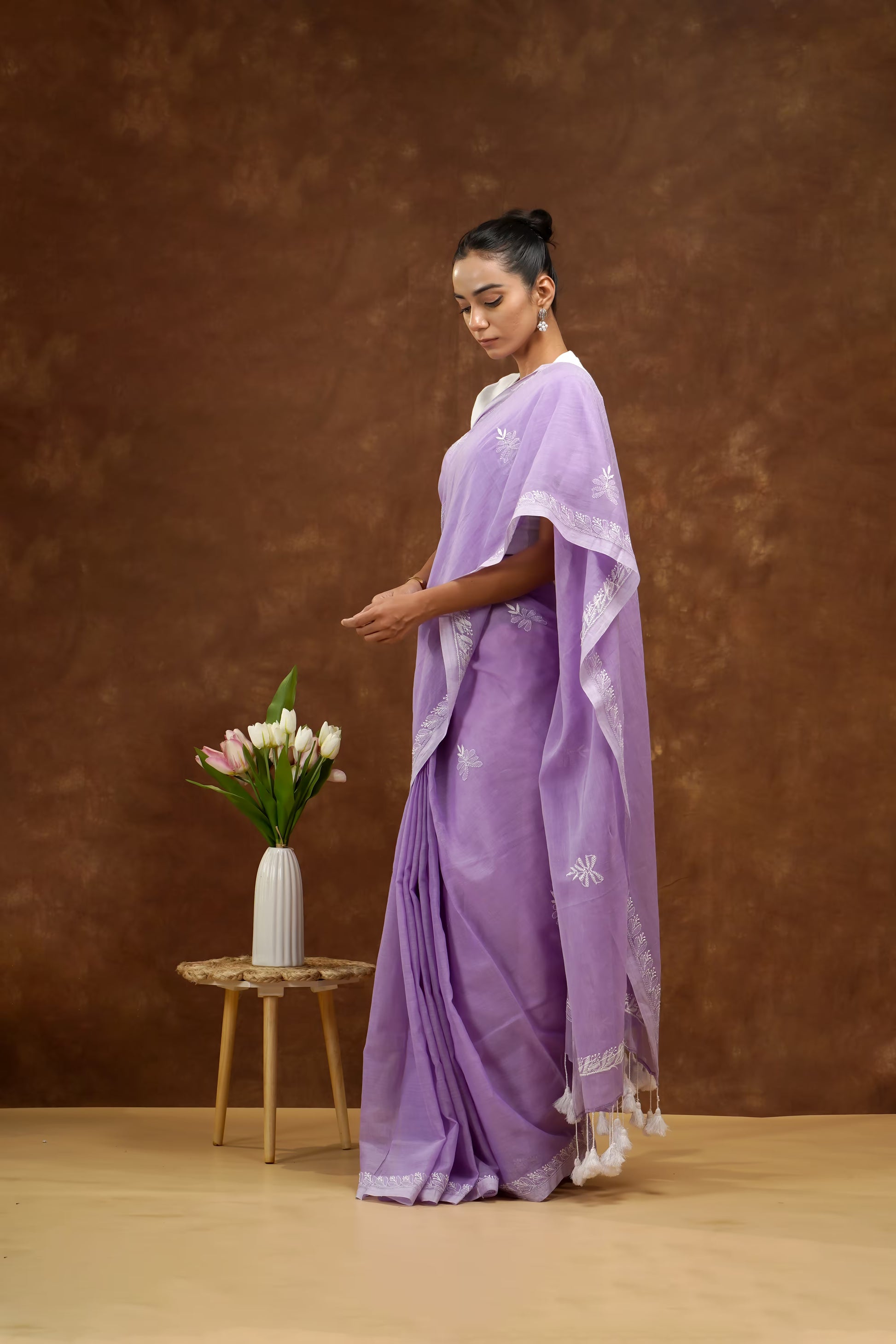 Woman in a purple saree standing next to a small table with flowers against a brown background