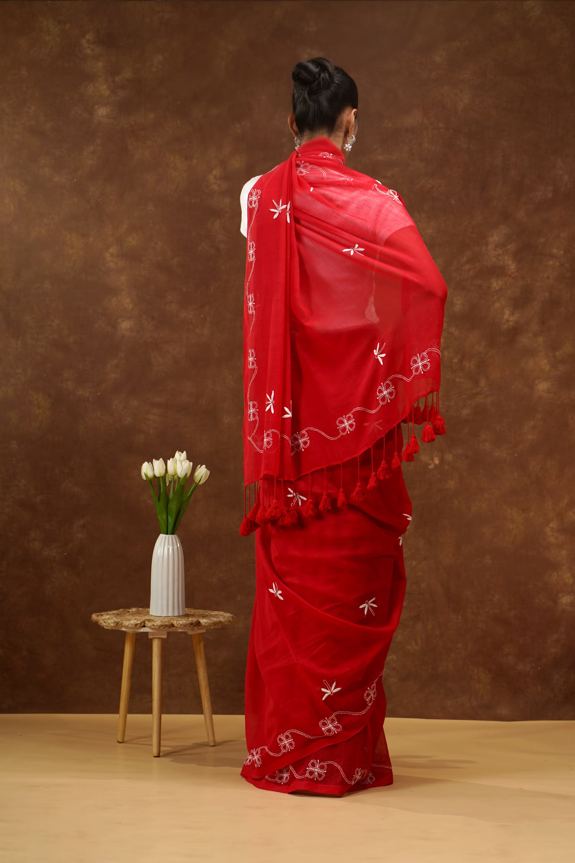 Person wearing a red saree with white patterns and tassels, standing against a brown background.