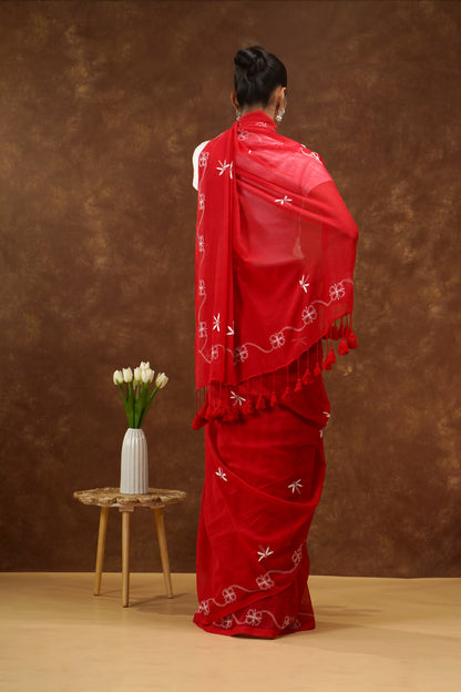 Person wearing a red saree with white patterns and tassels, standing against a brown background.