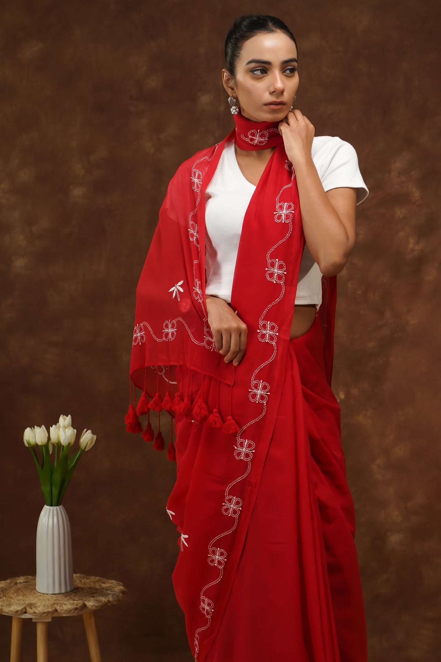 Woman wearing a red saree with white patterns and tassels against a brown background