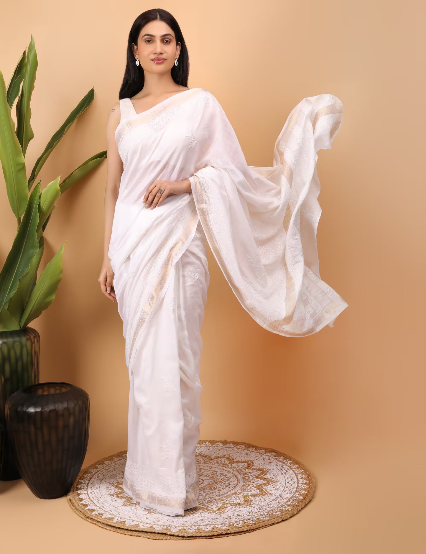 Woman in a white saree standing against a beige background with a plant to the left.