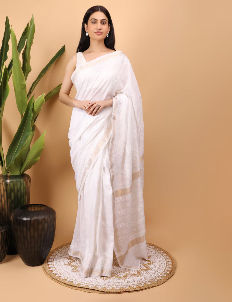 Woman in a white saree standing against a beige background with a plant to the left.