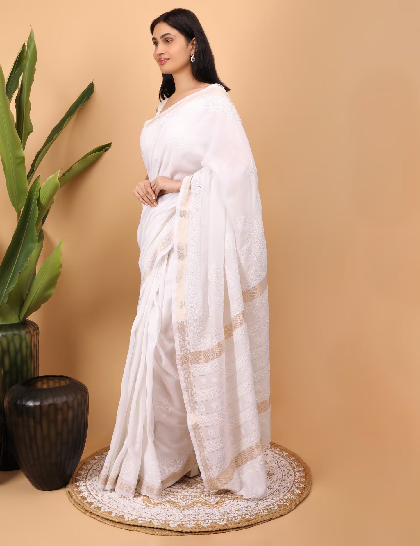 Woman in a white saree standing on a beige background with a plant to the left.