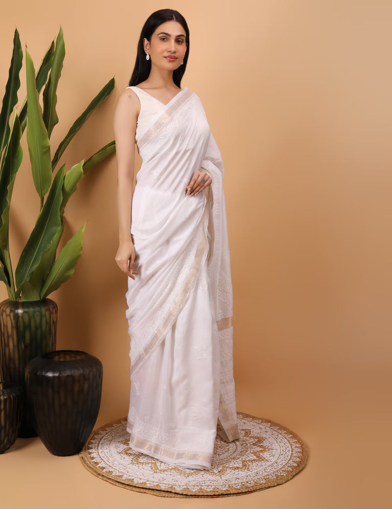 Woman in a white saree standing against a beige background with decorative elements.