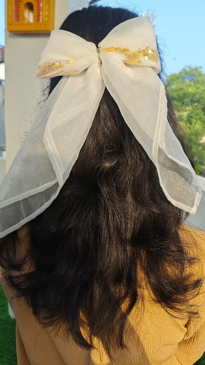 Person with dark hair wearing a large white bow with gold accents outdoors.