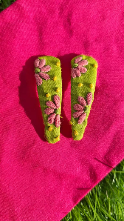 Two green clips with pink floral Embroidery on a pink fabric background