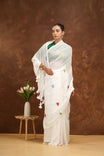 Woman in a white saree with floral patterns standing next to a small table with flowers against a brown background