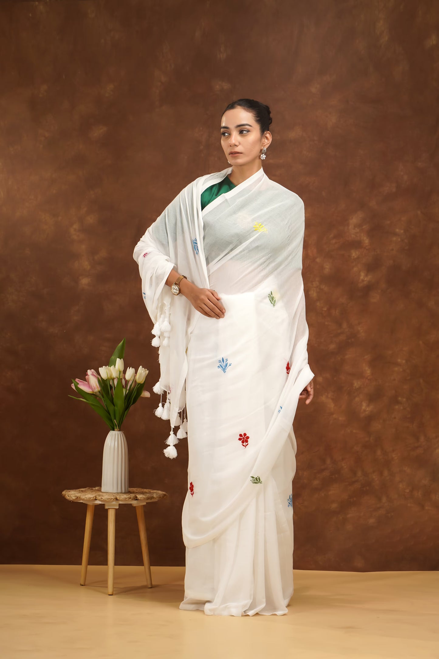 Woman in a white saree with floral patterns standing next to a small table with flowers against a brown background