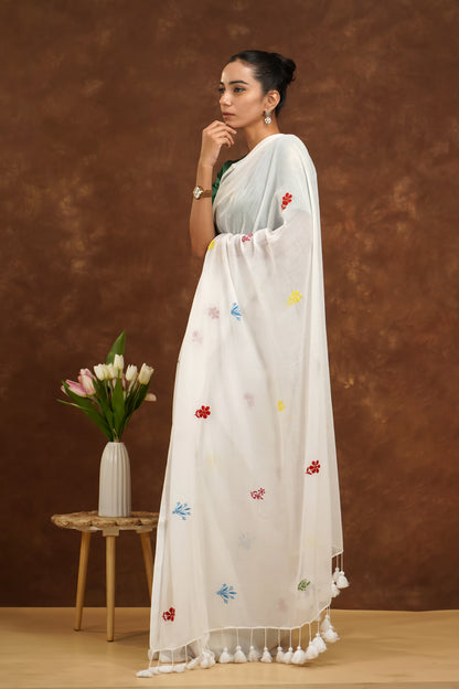 Woman in a white saree with colorful patterns standing against a brown background