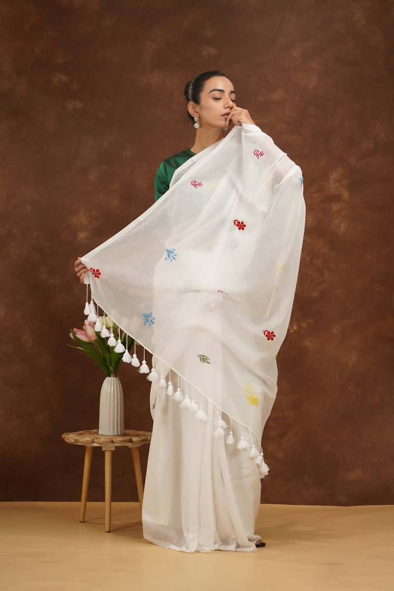 Woman in a white saree with colorful patterns and tassels against a brown background