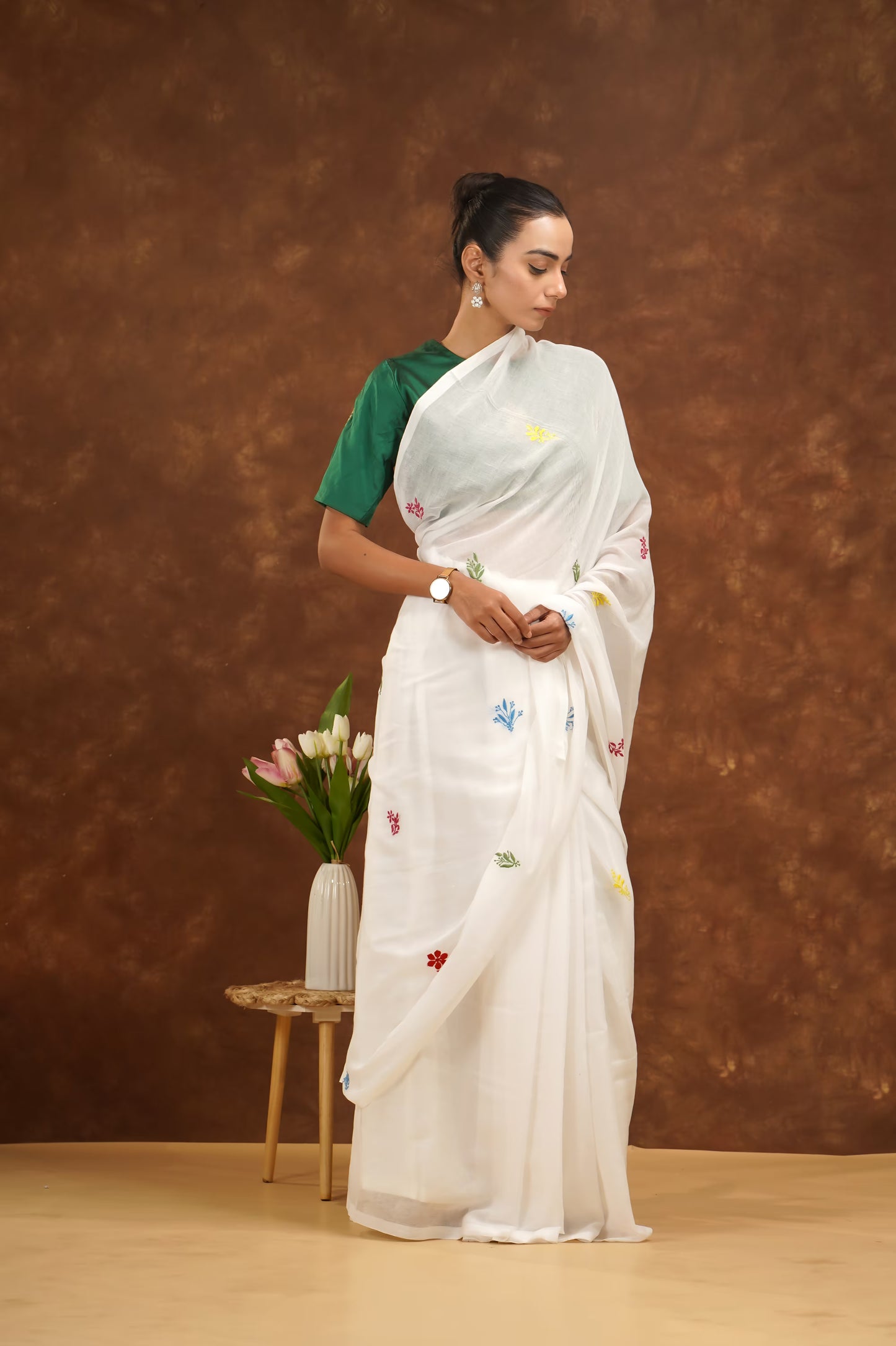 Woman in a white saree with floral patterns standing against a brown background