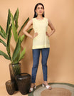 Woman wearing a light yellow Chikankari sleeveless top and blue jeans standing next to a plant on a beige background