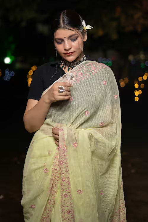 Woman wearing a light green saree with pink floral patterns against a dark background with bokeh lights.