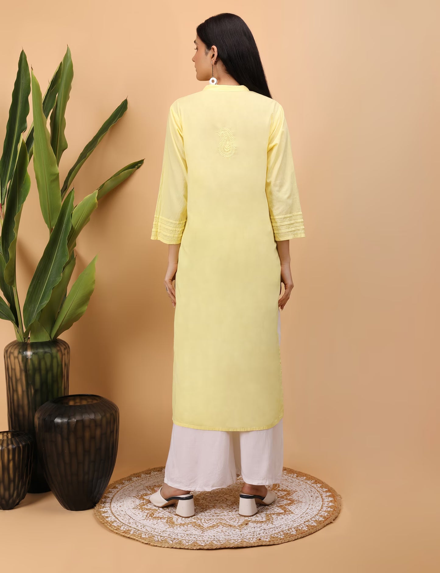 Woman wearing a yellow kurta with white pants standing in front of a beige wall with decorative vases.