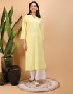 Woman wearing a yellow kurta with white pants standing against a beige wall with decorative vases and plants.