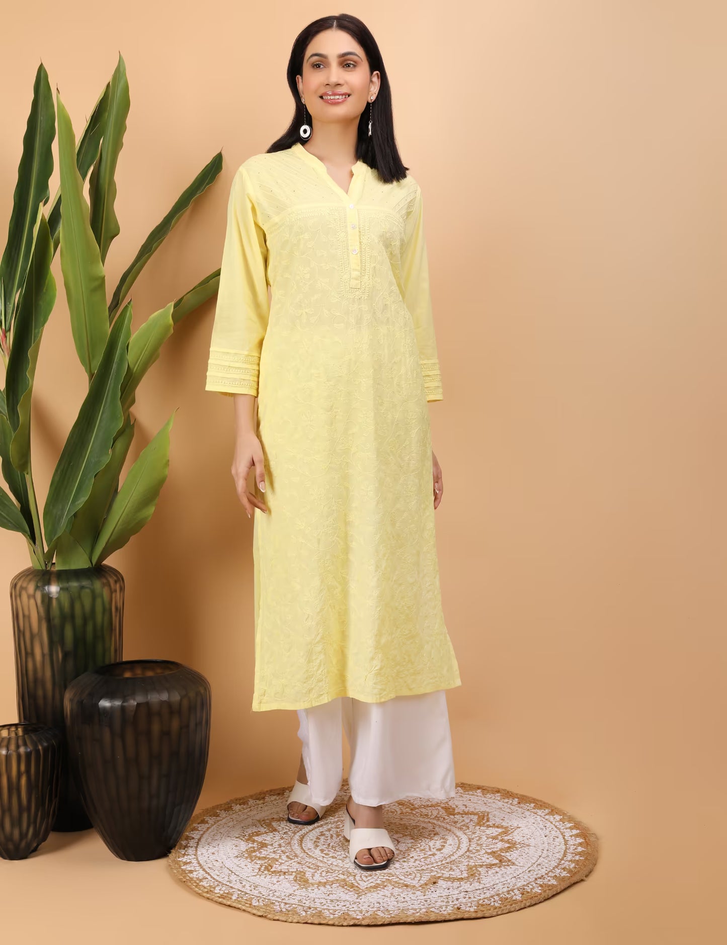 Woman wearing a yellow kurta with white pants standing against a beige wall with decorative vases and plants.