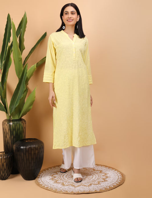 Woman wearing a yellow kurta with white pants standing against a beige wall with decorative vases and plants.