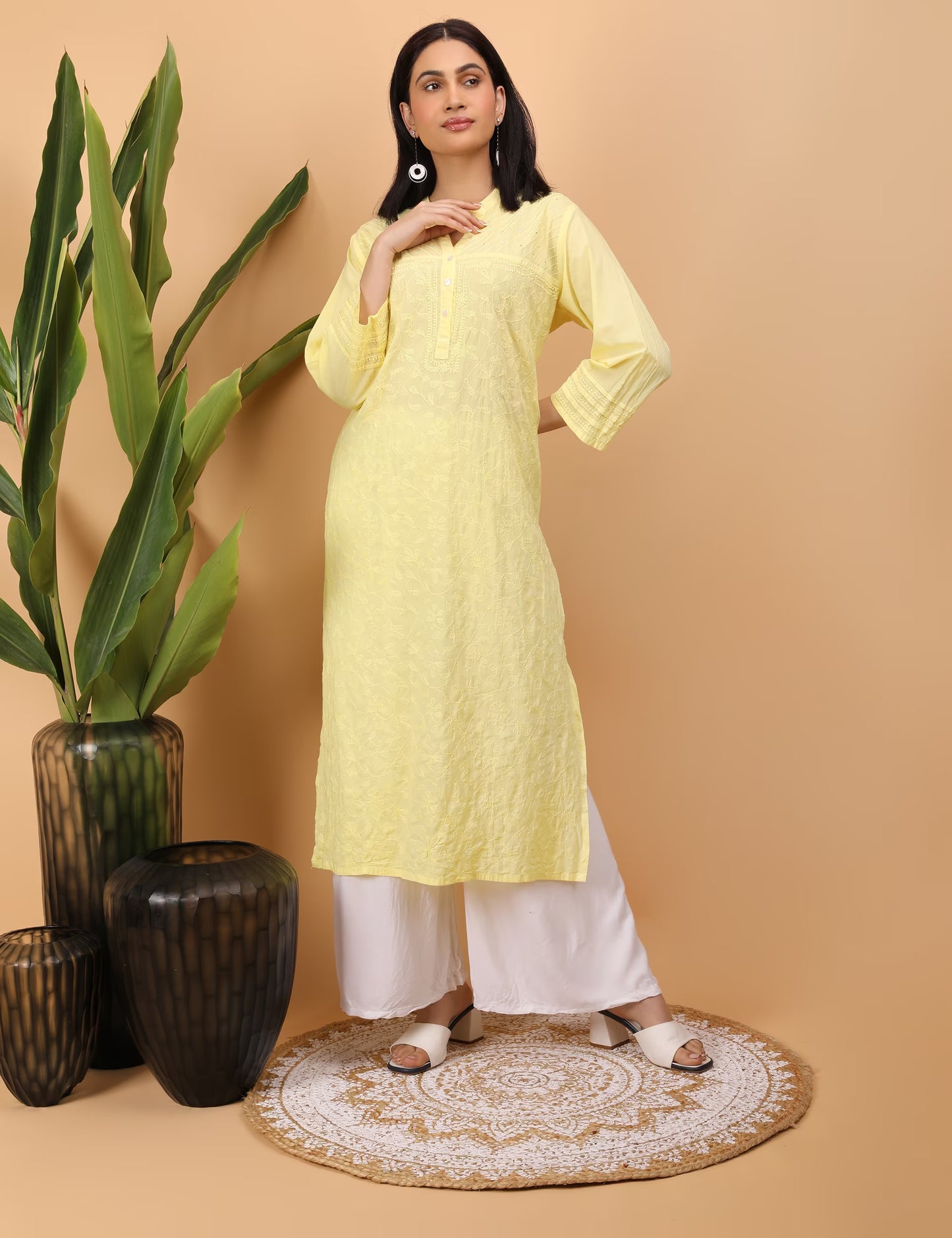 Woman in a yellow kurta with white pants standing next to decorative vases on a beige background