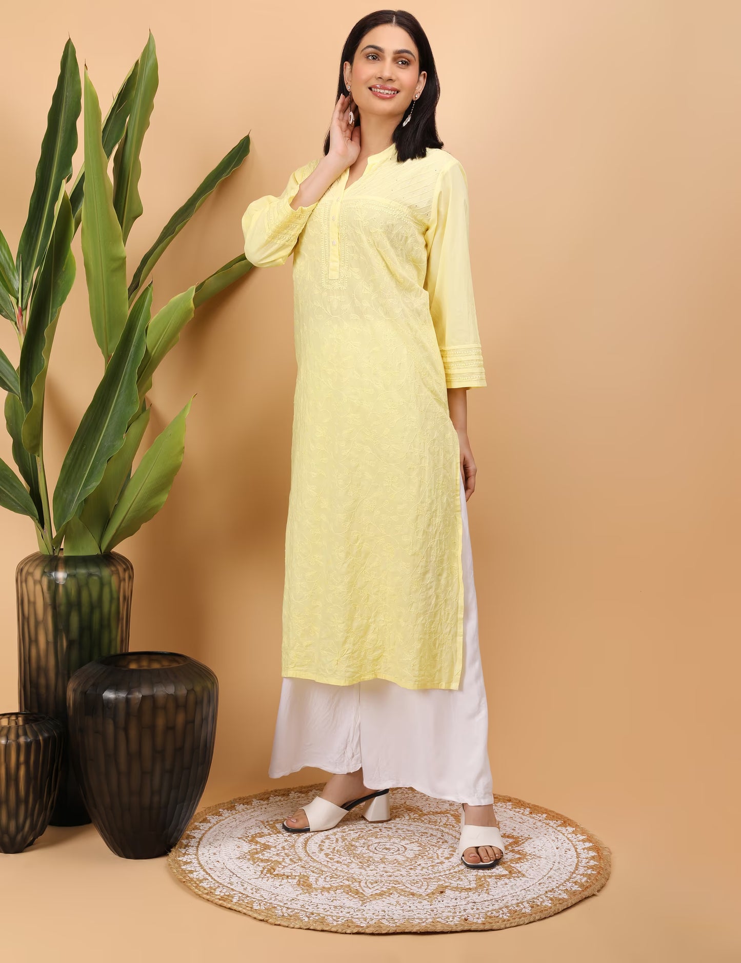 Woman wearing a yellow kurta with white pants standing next to decorative vases on a beige background