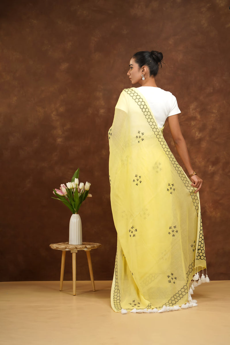 Woman in a yellow saree standing with a brown background and a small table with flowers.