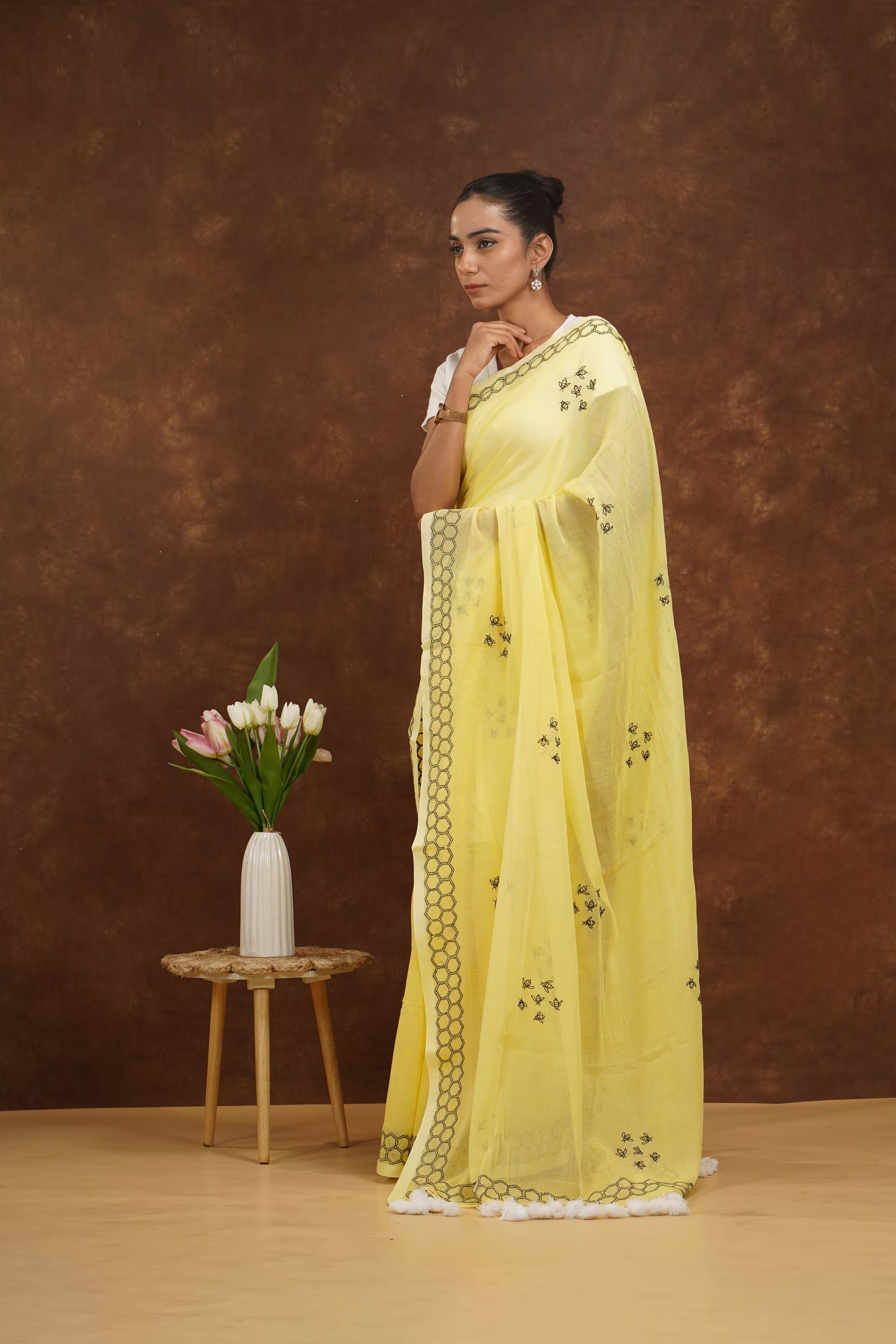 Woman wearing a yellow saree with a brown background and a small table with flowers.