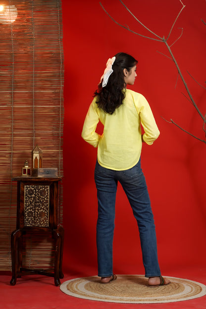 Woman in yellow top and blue jeans standing in front of a red wall with a small table and decorative items.