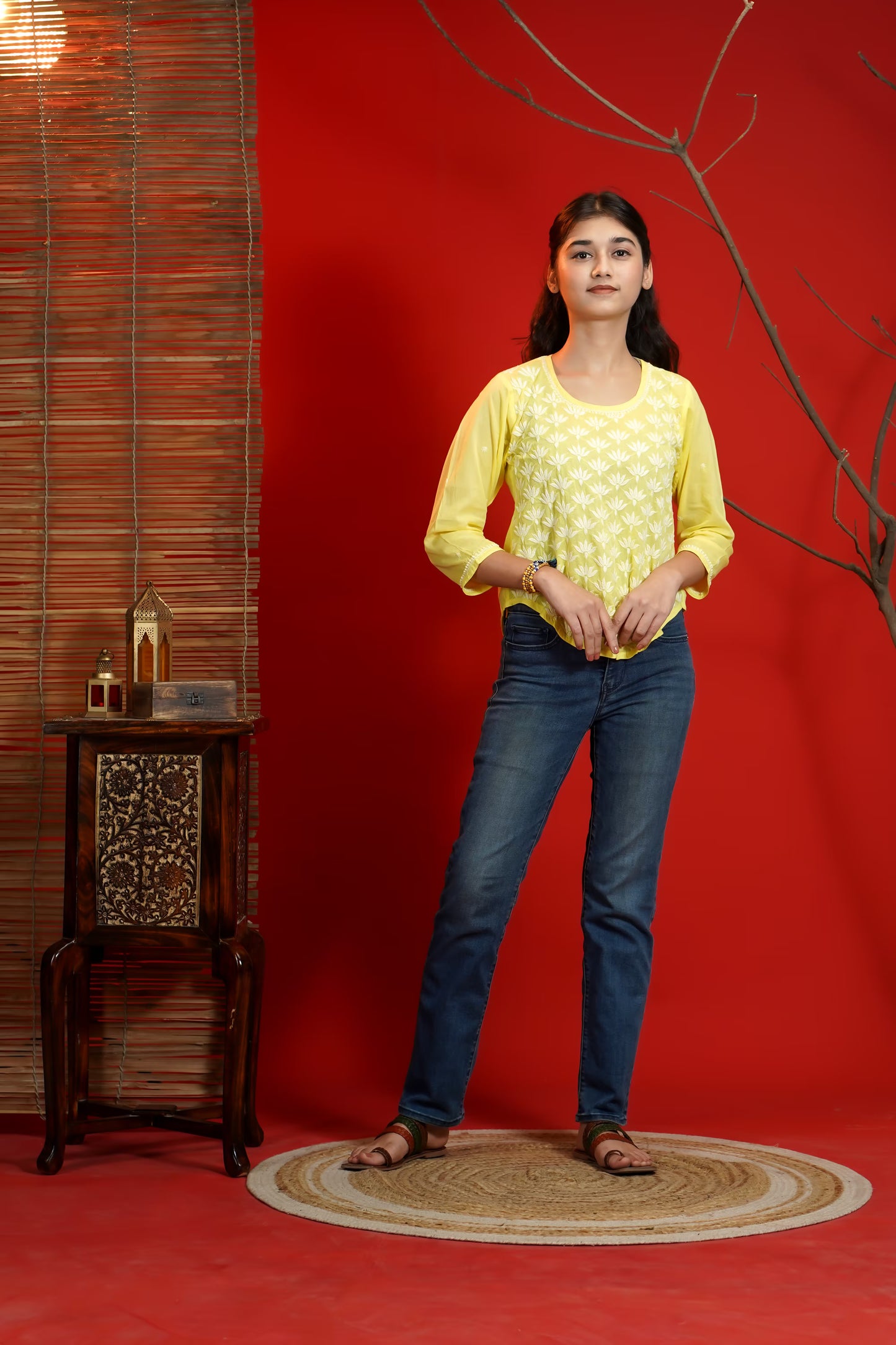 Woman in yellow top and blue jeans standing against a red wall with a wooden side table and plant.