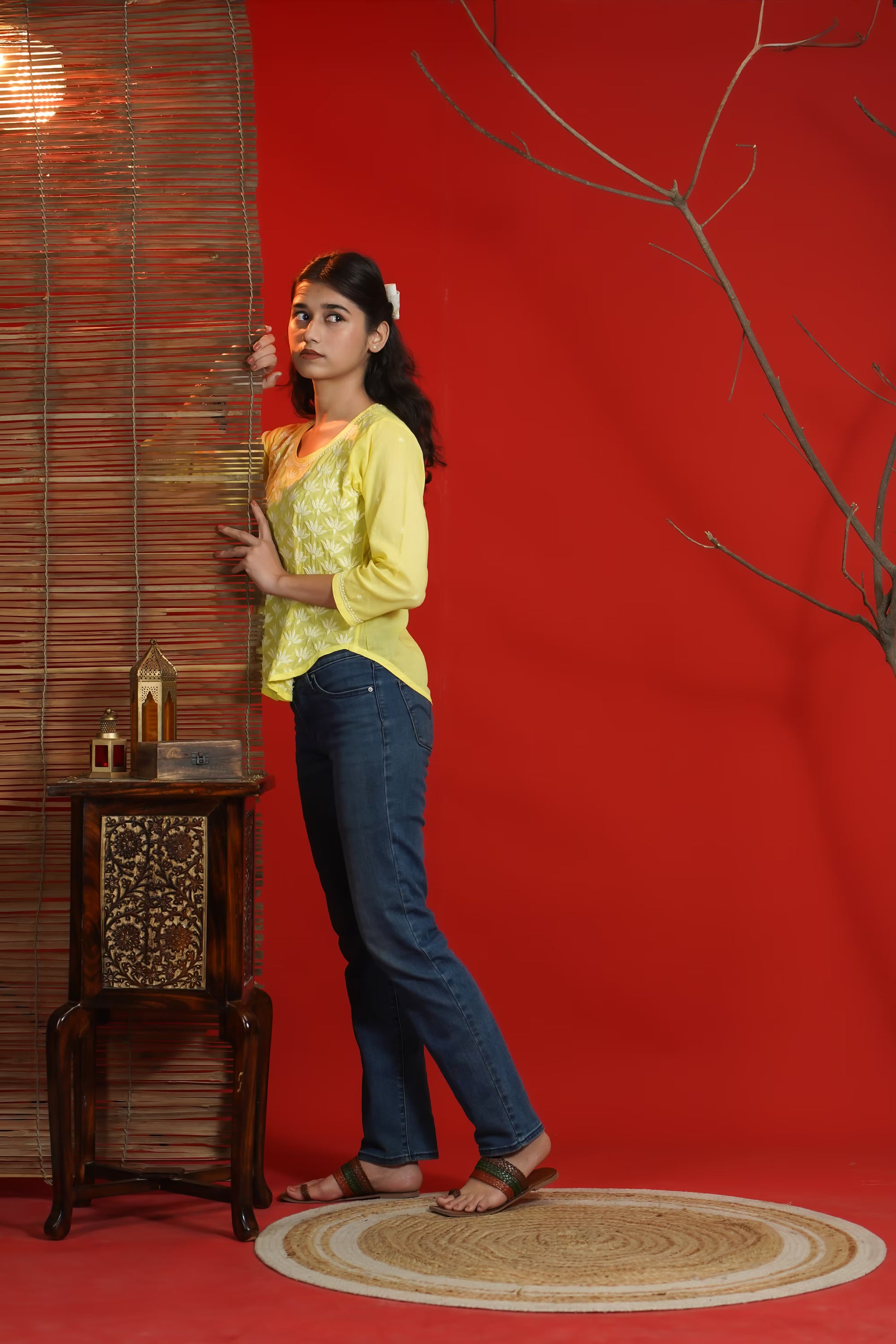 Woman in yellow top and blue jeans standing against a red wall with a wooden side table and decorative items.