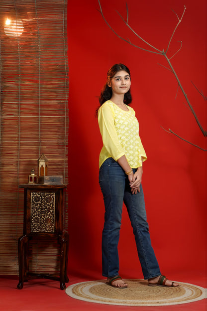 Woman in yellow top and blue jeans standing against a red wall with a decorative table and branch.