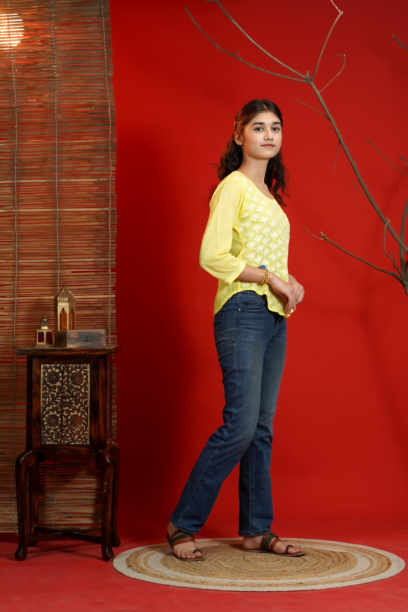 Woman in yellow top and blue jeans standing against a red wall with a small table and decorative items.