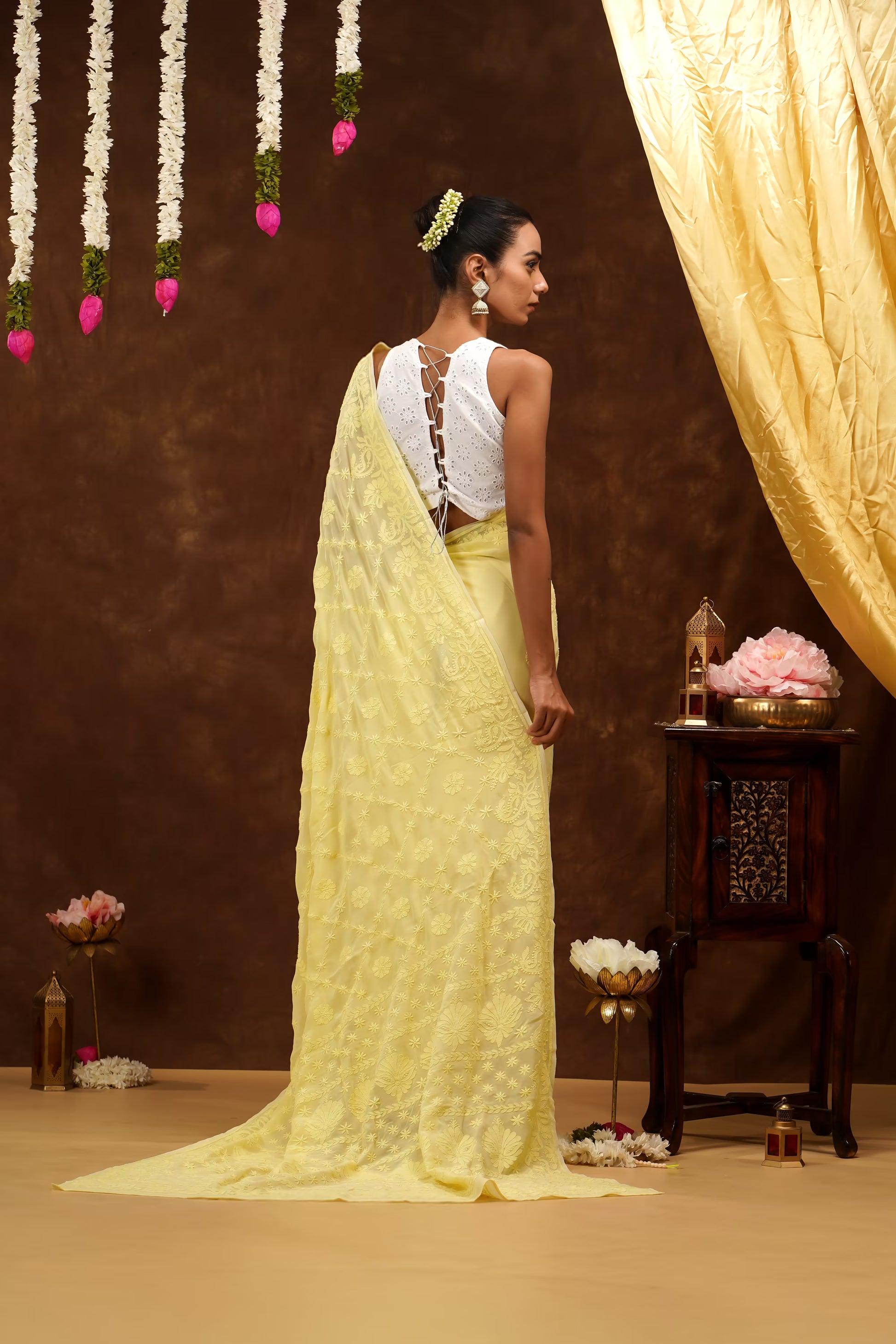 Woman in a yellow saree standing in a decorated room with flowers and a curtain.