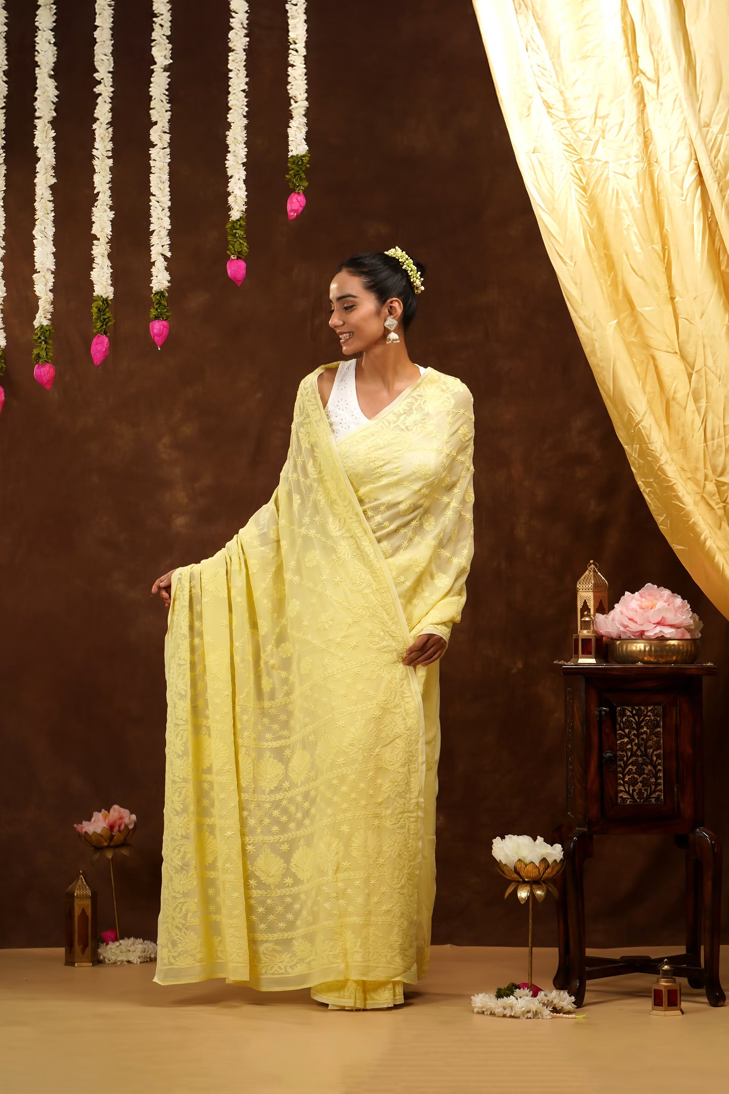 Woman in a yellow saree standing in a decorated room with flowers and curtains.