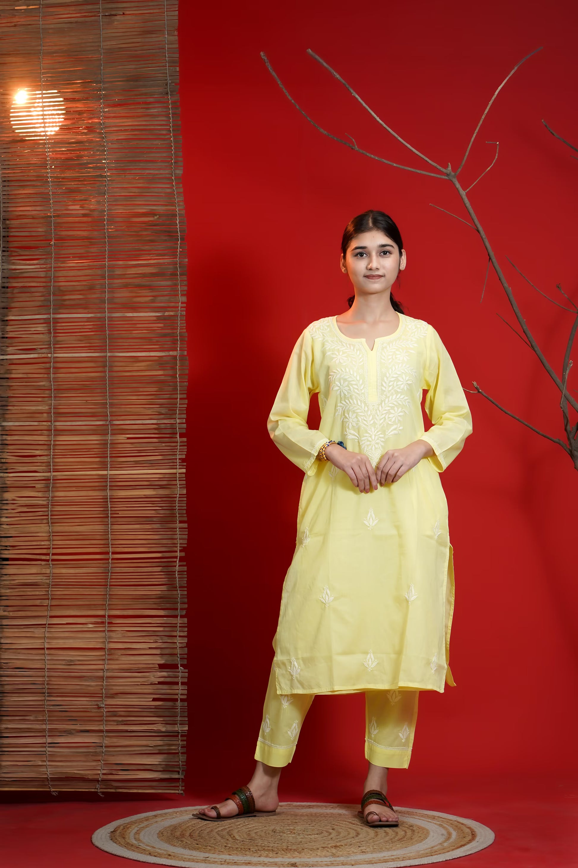 Woman in a yellow traditional outfit standing against a red wall with a textured wall and branch in the background.