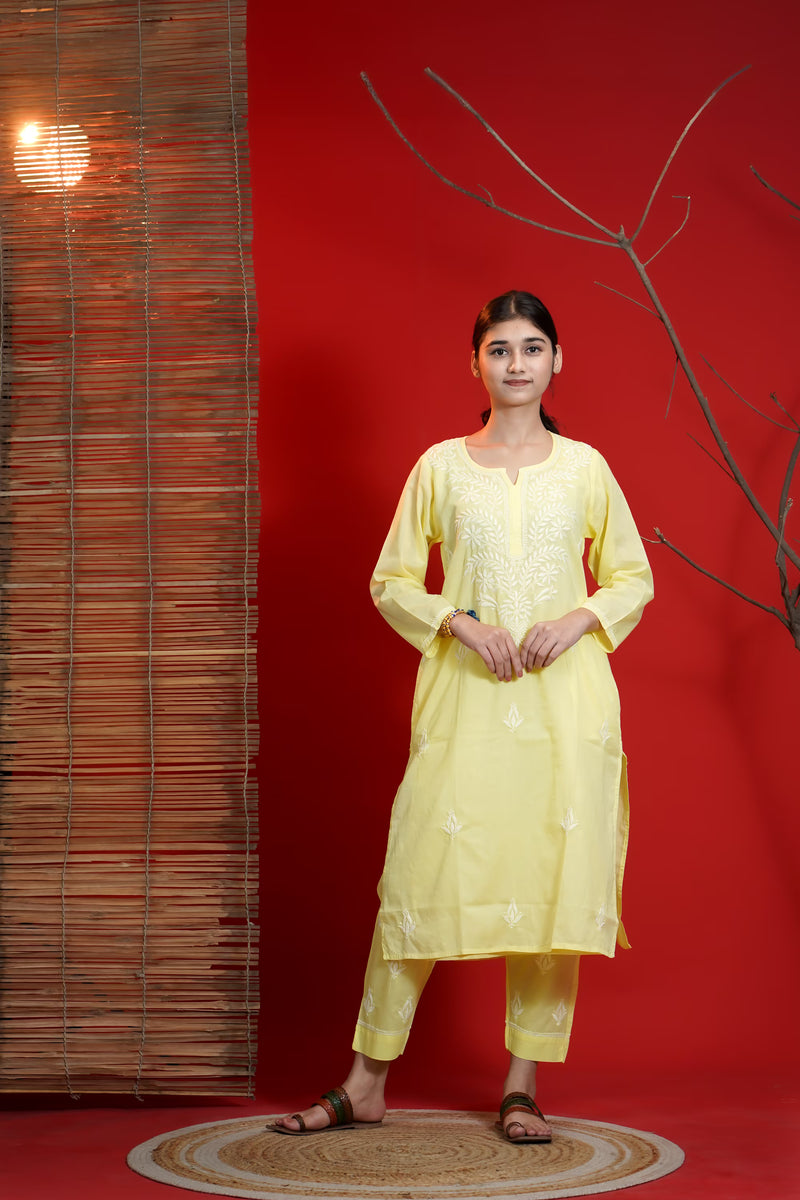 Woman in a yellow traditional outfit standing against a red wall with a textured wall and branch in the background.